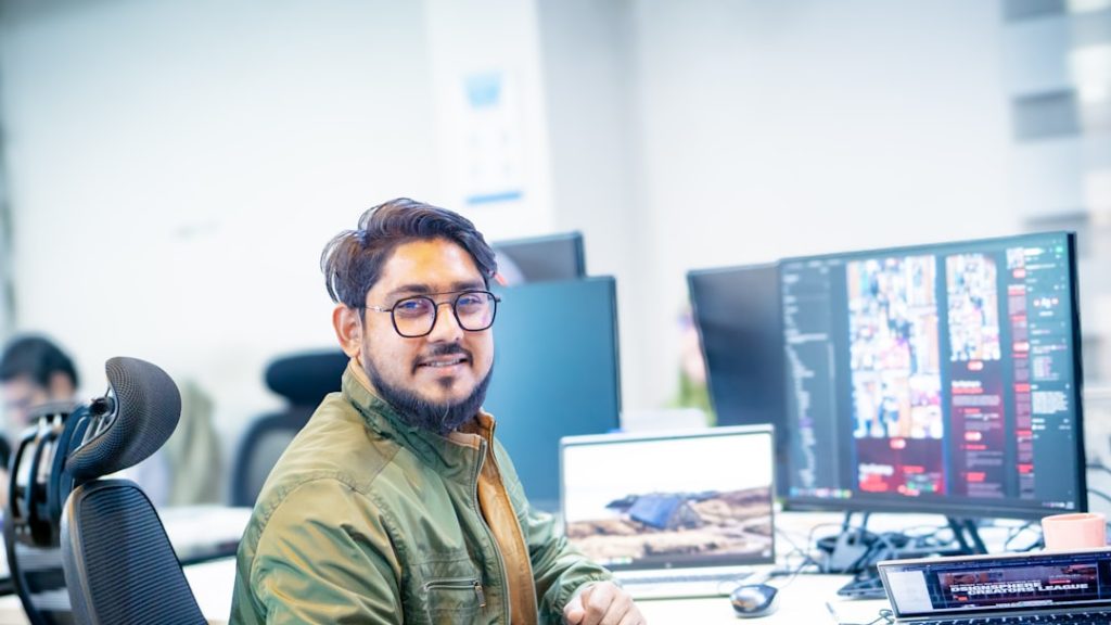 A smiling man wearing glasses and a green jacket works at his computer in a modern office filled with screens, creating a vibrant and productive tech workspace atmosphere.